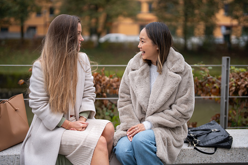 A mid adult Caucasian woman and her Asian friend are seated on a park bench, deep in conversation, enjoying a crisp autumn day in the city.