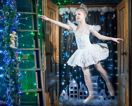 Cute pre-adolescent girl ballet dancer Is dressed in white beautiful tutu. She is flying in ballet pose, smiling and looking away. Studio shoot at the doorway with a cityscape and a string light as a background
