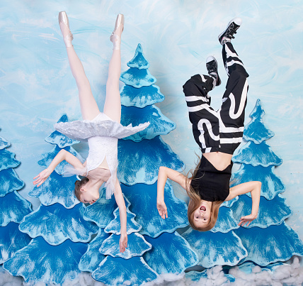 Two funny girls (ballerina and her friend) are flying upside and waving hands on blue background with fake spruce trees. The preteen girls are screaming and looking at the camera. Studio shooting, upside down picture