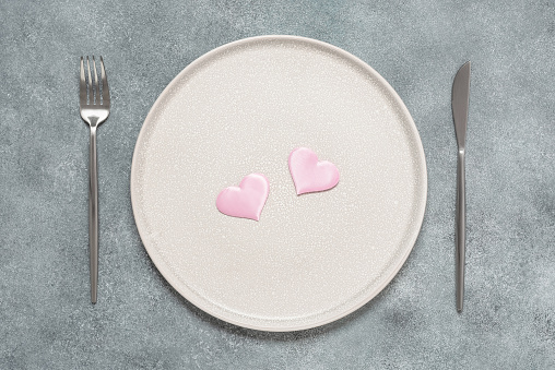 Minimal table setting for Valentine's Day. Two pink hearts on a beige plate, fork, knife. Top view, flat lay. Gray grunge background.