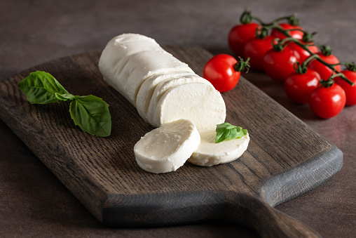 Mozzarella cheese sliced on a wooden cutting board with basil leaves and cherry tomatoes, side view, dark old background.