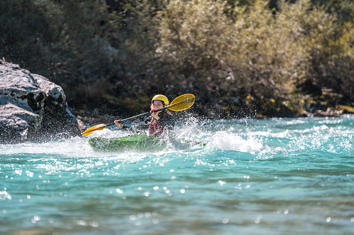 A young Caucasian woman showcases her paddling skills in a green kayak on a river, equipped with a yellow helmet and red life vest, embodying the spirit of water sports.