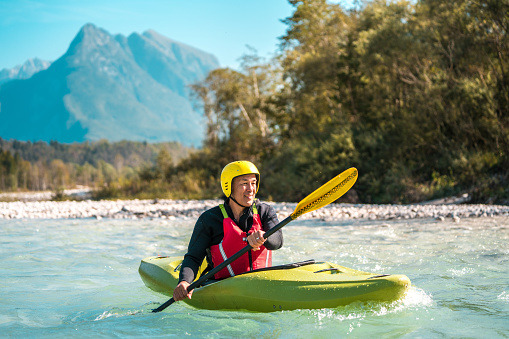 An Asian male kayaker navigates the rapids of a mountain river, equipped with a yellow helmet and paddle in a vibrant green kayak.