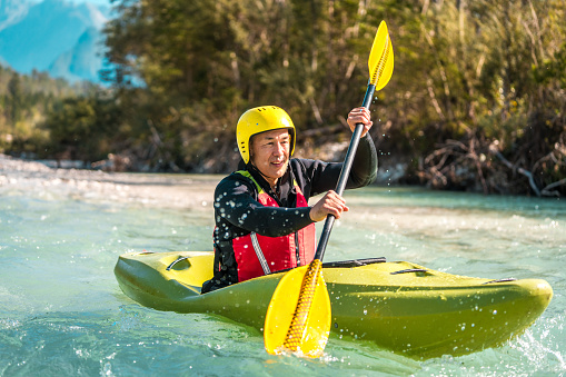 Mature Asian male exhibiting active lifestyle, paddling a green kayak, surrounded by lush forest and mountains.