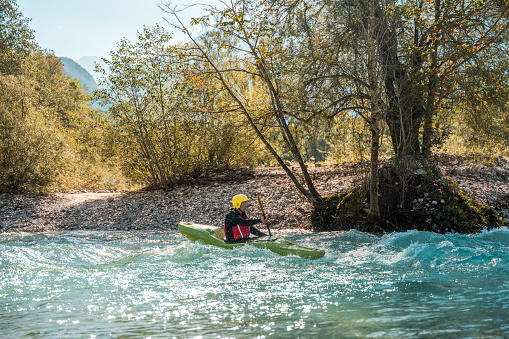 Mid adult male kayaker in a yellow helmet and red life jacket skillfully maneuvers through the turbulent waters of a scenic river, surrounded by lush greenery.