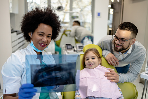 In a dental clinic, a dentist in a coat and gloves shows x-ray images to a young patient and her father, emphasizing preventive care and the importance of family involvement in dental health