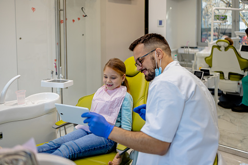 In a dentist's office, a child looks at a digital tablet held by the dentist, showcasing the use of wireless technology in pediatric dental examinations and care