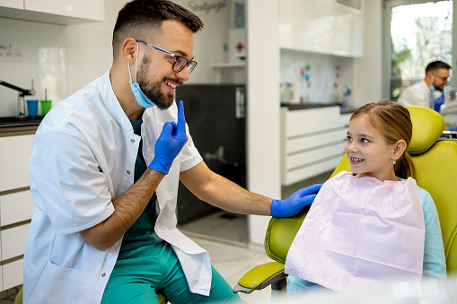 A small girl, looking away with a positive emotion, sits in an office chair while a dentist in a protective face mask checks her teeth, ensuring a pleasant experience