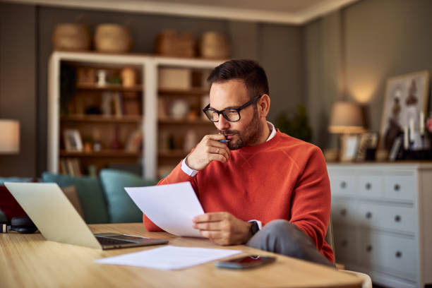 istock A concentrated male college professor with glasses grading exam papers from students while sitting in his office in front of a laptop. 1927152018