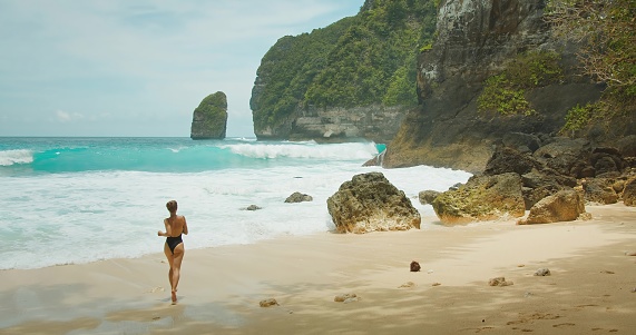 Woman contemplates the vast sea. Female in swimsuit running on sandy beach, powerful turquoise waves crashing against the rocky cliffs, a moment of solitude amidst the majesty of nature.