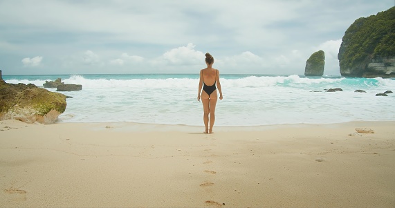 Adult tanned girl in swimsuit stands on sandy beach and watches the ocean. Sea waves splash. Rear view of brunette woman looking at seascape. The concept of a romantic tropical vacation.