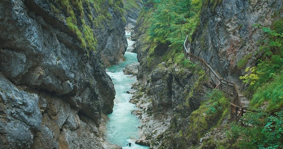 Hiking trail in gorge with mountain fast flowing river at rainy day. Narrow wooden staircase along the canyon in Austria. Autumn cloudy foggy nature. Lammerklamm ravine.