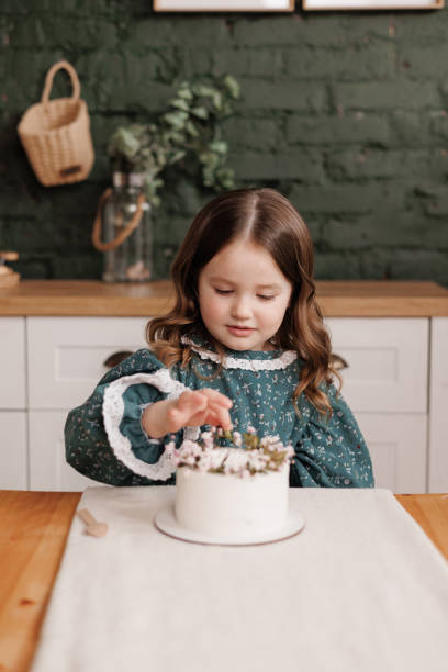 愛らしい小さな子供の女の子は、自宅の誕生日パーティーでテーブルの上に飾られた花の名前のケーキでお祝いのドレスを着ています。幸せな陽気な子供は、屋内でデザートに白いクリーム� - preschooler caucasian one person part of ストックフォトと画像