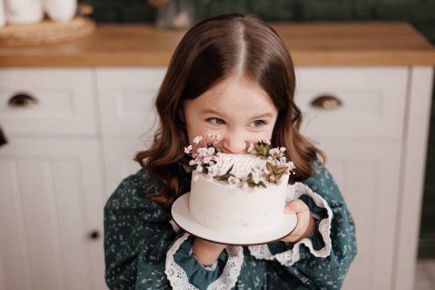 愛らしい小さな子供の女の子はお祝いのドレスを着て、誕生日パーティーで飾られた花の名前のケーキを一口食べます。幸せな笑顔の子供が自宅のキッチンでデザートに白いクリームをなめ� - preschooler caucasian one person part of ストックフォトと画像