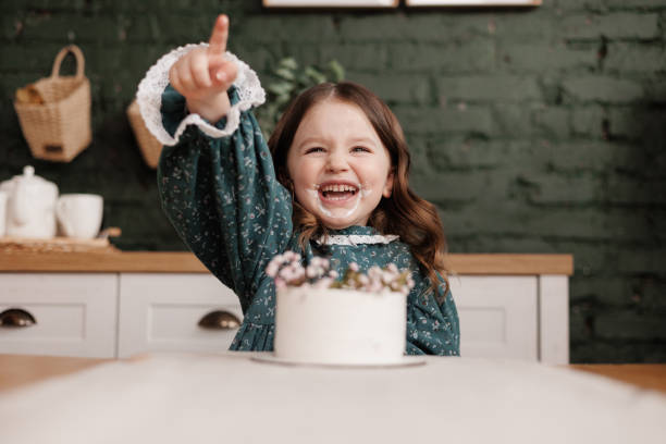 adorable little child girl wears festive dress takes a bite out of a decorated flowers name cake at a birthday party. happy smiling kid licks white cream from her dirty face and shows thumb up indoors - preschooler caucasian one person part of ストックフォトと画像