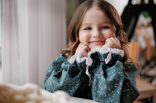 Adorable little girl with curly hair wears festive rural style dress is smiling, looking at camera, sitting by the table and supports her chin with hand at home. Happy childhood concept. Children day.