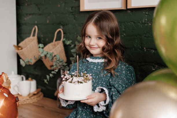 adorable little child girl 4-5 years hold white birthday cake decorated with flowers and trying to blow burning candle. kid wears festive stylish retro dress is ready to her birthday party at home - cute 4 5 years little girls party стоковые фото и изображения