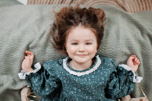 child birthday celebration. top view of happy little girl 4-5 years wears stylish rustic style dress is lying on bed, smiling and looking at camera. - preschool 2 3 years preschooler little girls - fotografias e filmes do acervo