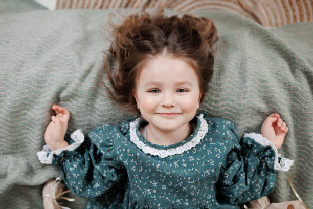 child birthday celebration. top view of happy little girl 4-5 years wears stylish rustic style dress is lying on bed, smiling and looking at camera. - cute 4 5 years little girls party стоковые фо�то и изображения