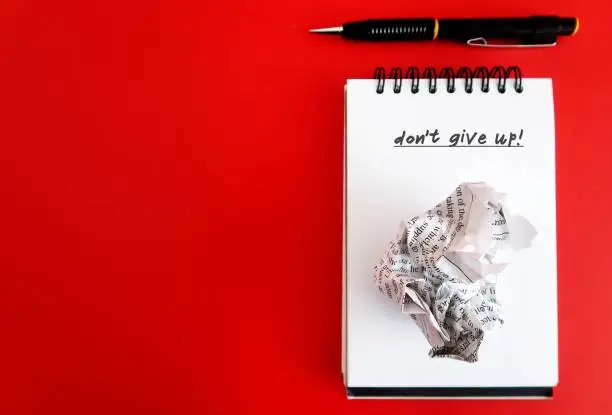Pen and crumpled paper on notebook with text DO NOT GIVE UP on red copy space background, concept of self motivated to keep on writing, do not give up even the words are not flowing ,overcome writers block