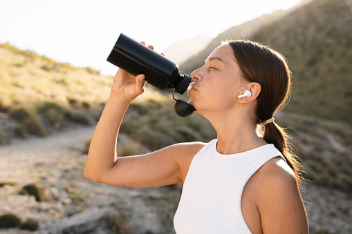 Rehydration. Female caucasian athlete drinking water from sporty bottle after tedious training jogging running. Water balance with electrolytes. Yoga outdoors in the mountains