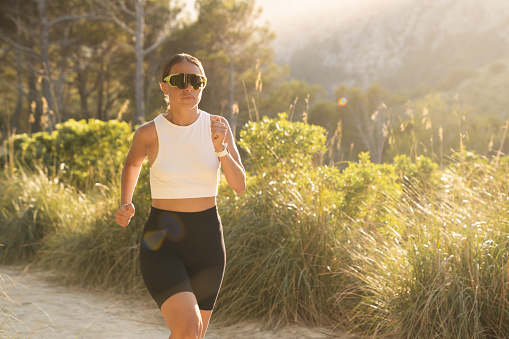 Closeup cropped image of active young caucasian female athlete running doing sportive exercises while jogging in the park forest outdoors. Training in fitness clothes with sunglasses.