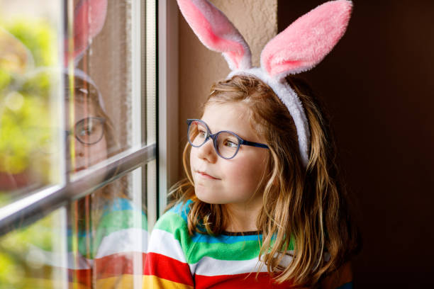portrait of a happy little girl with bunny ears looking outside sitting by a window. easter holiday. adorable child happy about holiday. - easter easter bunny fun humor stock-fotos und bilder