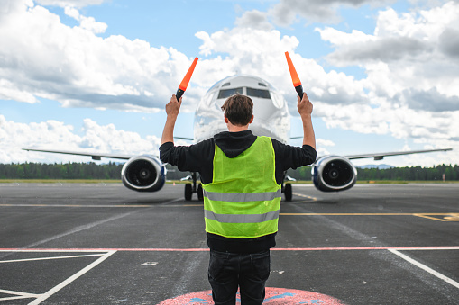 Caucasian airport worker marshalling an aircraft. Wearing a reflective vest and holding signal lights for marshalling.