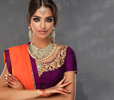 Young woman dressed in a traditional Indian suit lehenga choli, purple blouse with gilded decoration, posh jewelry set  of big earrings, bright necklace, head adornment (tikka) and henna tatto on the hands.Indian style.