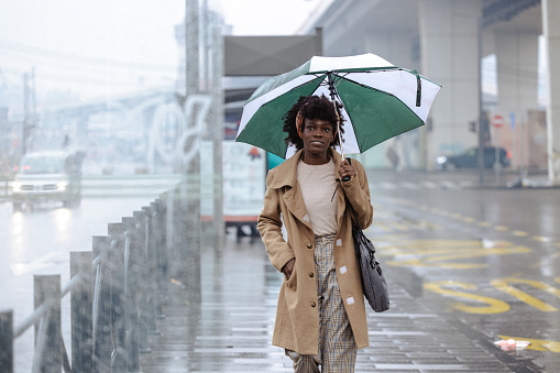 Shot of a smiling African American woman on a rainy day with umbrella waiting for a bus on station