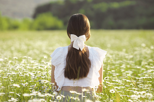 Ponytail faceless woman hairstyle with white bow clip on long brown hair. A young girl from the back sits in a field of daisies and enjoys freedom and calm. Mental health. Chamomile field landscape.