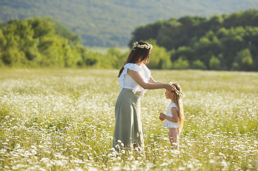 Lifestyle mother putting chamomile wreath on daughters hair. Mothers day concept. Slavic tradition wreaths from wild daisy flowers.