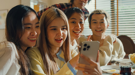 Group of young Asian people watching smartphone and having fun sitting at dining table at home. Multicultural friends enjoying spending together college house party concept.
