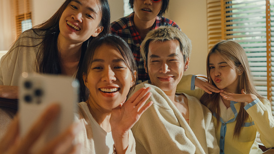 Closeup group of young Asian people using mobile phone taking selfie and having fun sitting at dining table at home. Multicultural friends enjoying spending together college house party concept.