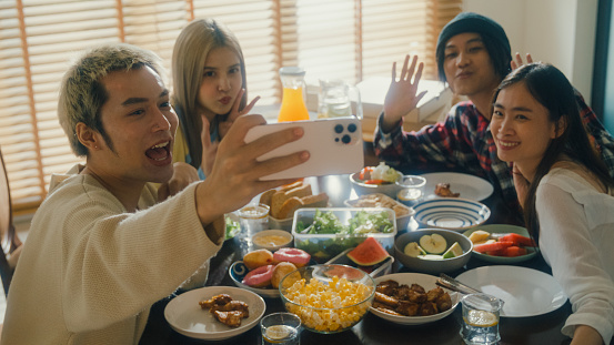 Group of young Asian people using mobile phone taking selfie and having fun sitting at dining table at home. Multicultural friends enjoying spending together college house party concept.
