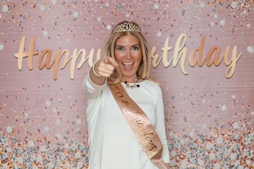 Cheerful blond woman in a white t-shirt, wearing a crown with number 50 having fun in a studio with Happy Birthday background and pointing at camera.
