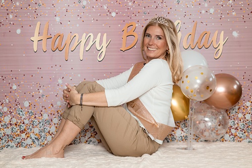 Cheerful blond woman in white and brown casual clothes  and a crown with number 50 on the head, sitting on the blanket on the floor and posing in front of the background with Happy Birthday sign while looking at camera.