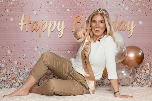 Cheerful blond woman in white and brown casual clothes  and a crown with number 50 on the head, sitting on the blanket on the floor and posing in front of the background with Happy Birthday sign while looking and pointing at camera.