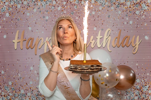 Adult blond woman in a white t-shirt holding a cake with a candle and looking up while making a wish. Happy Birthday studio background.