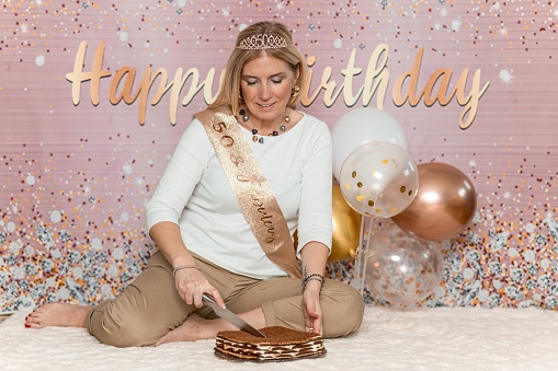 Adult woman cutting a birthday cake on the white blanket. Balloons and Happy Birthday background behind her.