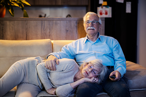 A senior man is watching tv late and his wife is relaxing and napping in his lap.