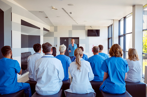 Back view of group of doctors and nurses sitting in a conference room and listening to female speaker. Mature woman giving a speech on seminar for medical staff.