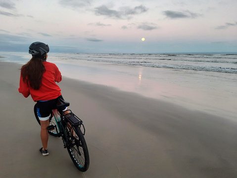 Young woman standing with a bicycle on a beach looking at the moon