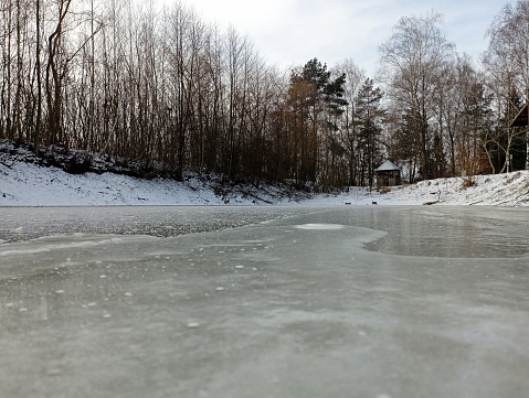 The lake is completely covered with thick ice. Winter landscape on a small pond. There is a wooden gazebo on the far bank of the pond. Trees grow around the pond.