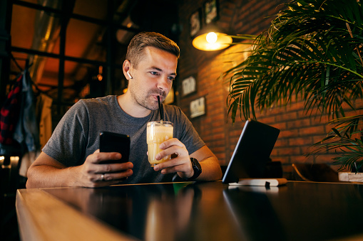 A trendy man is sitting at restaurant and drinking a juice while using a phone and looking at online menu on tablet.