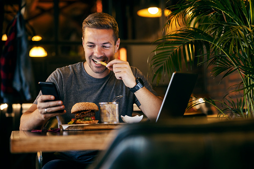 A casual businessman is sitting at restaurant and eating his lunch while smiling at the phone and reading e-mails.