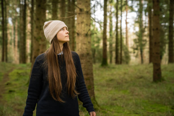 A white woman, with brown hair and a veis hat, in an Irish forest with green and brown tones through the trees