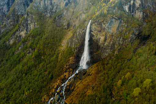 High-angle drone panoramic photo of tall and powerful waterfall falling into the crystal blue river in beautiful valley surrounded by green mountain peaks in Western Norway, Scandinavia