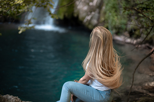 Young woman sitting in nature next to waterfall on windy day, her hair is waving