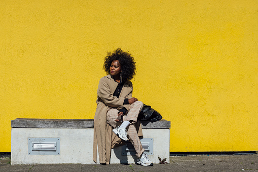 Full length front view portrait of a mature stylish woman, she is sitting in front of a yellow wall background in the city, she is holding a bag and is wearing fashionable clothing while using a smartphone. She is looking away from the camera.